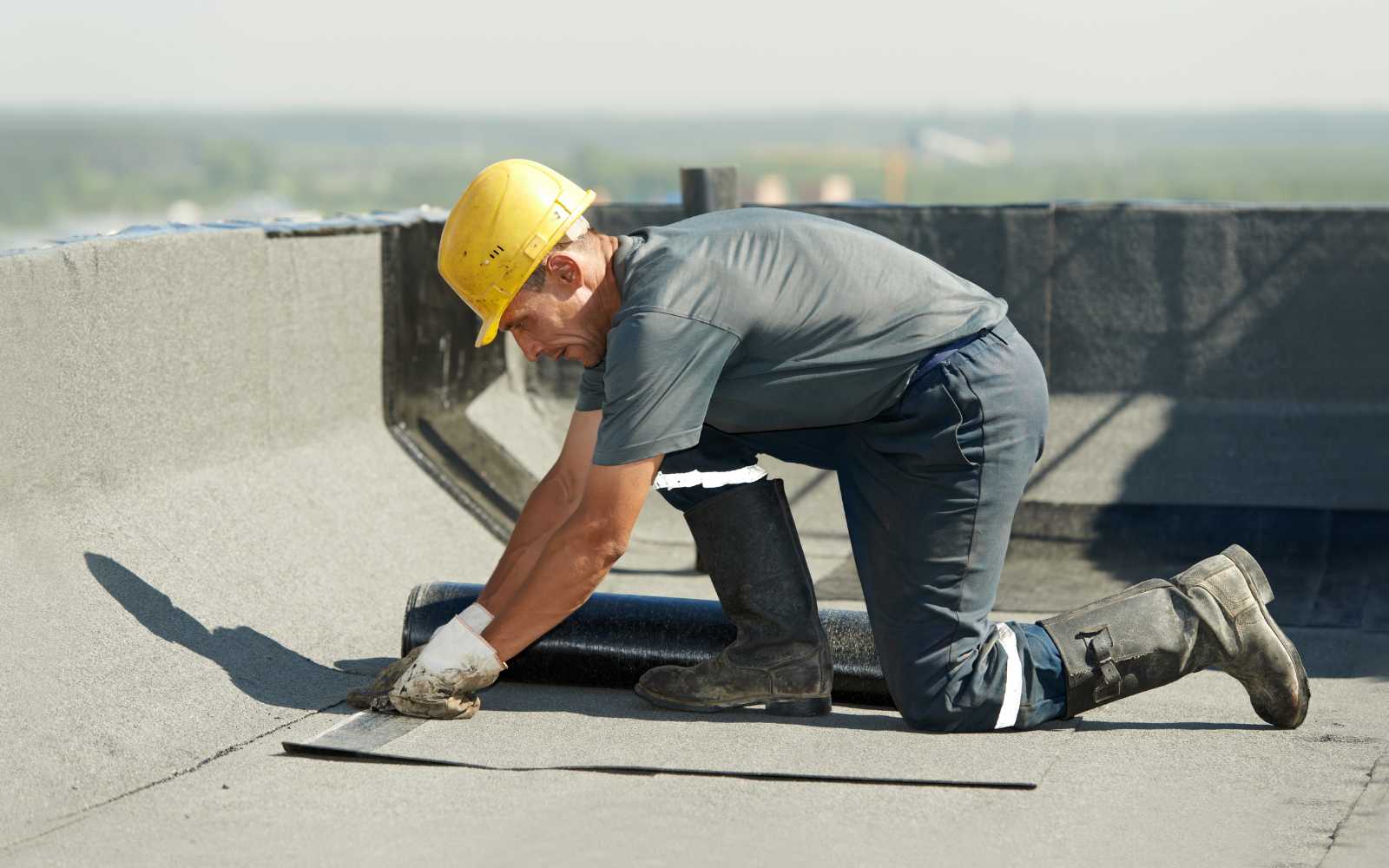 A worker in a yellow hard hat is installing a roll of dark roofing material on a flat roof.
